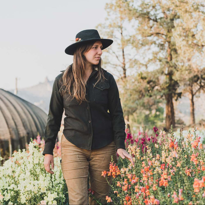 Woman in a black jacket and hat standing among flowers with trees in the background