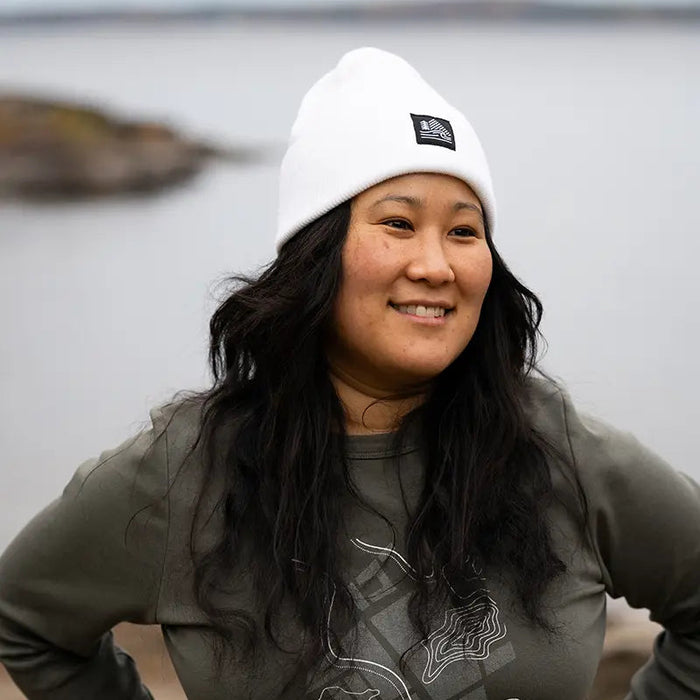 Woman wearing a white beanie and gray sweatshirt with a logo, standing by a body of water.