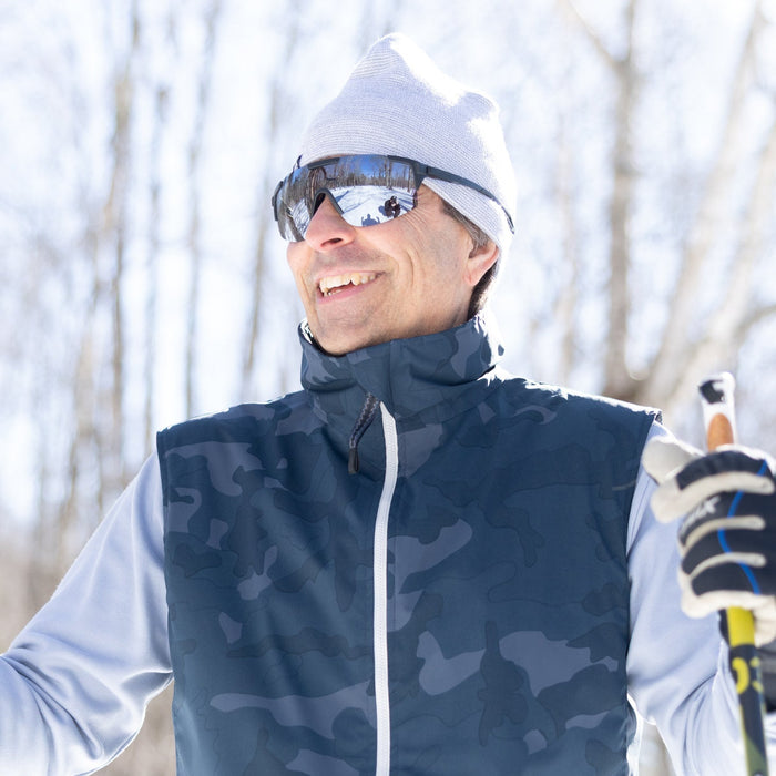 Man skiing in a snowy landscape wearing a blue camouflage vest, white helmet, and goggles.