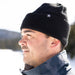 Man wearing a black knit beanie and blue jacket outdoors with a blurred natural background