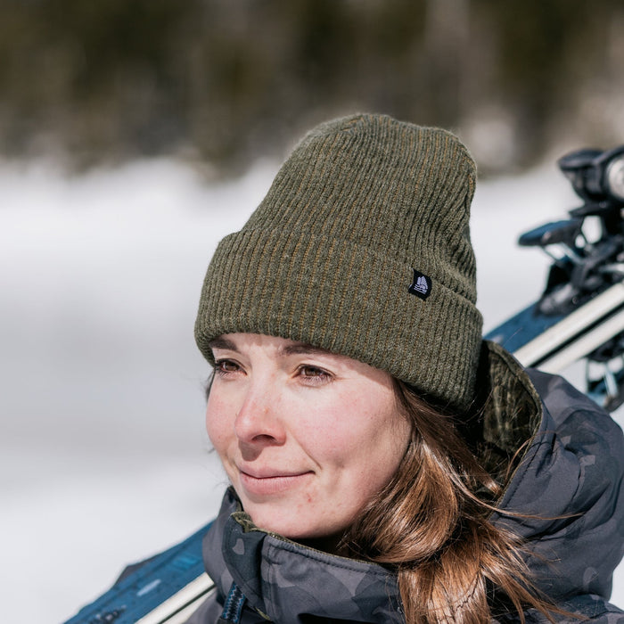 Person wearing a green beanie and winter coat in a snowy landscape with trees in the background.