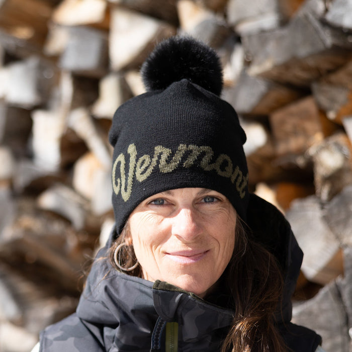 Person wearing a black beanie with 'Vermont' text in front of stacked firewood.