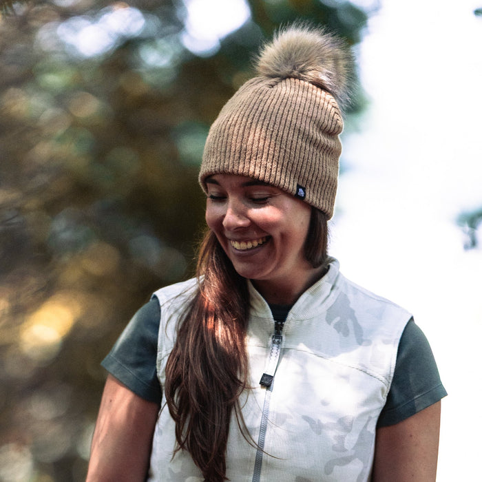 Woman wearing a beige beanie with a pom-pom and a white vest outdoors.