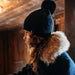Person wearing a dark coat with fur hood and a blue knit beanie in a warm, wooden interior.