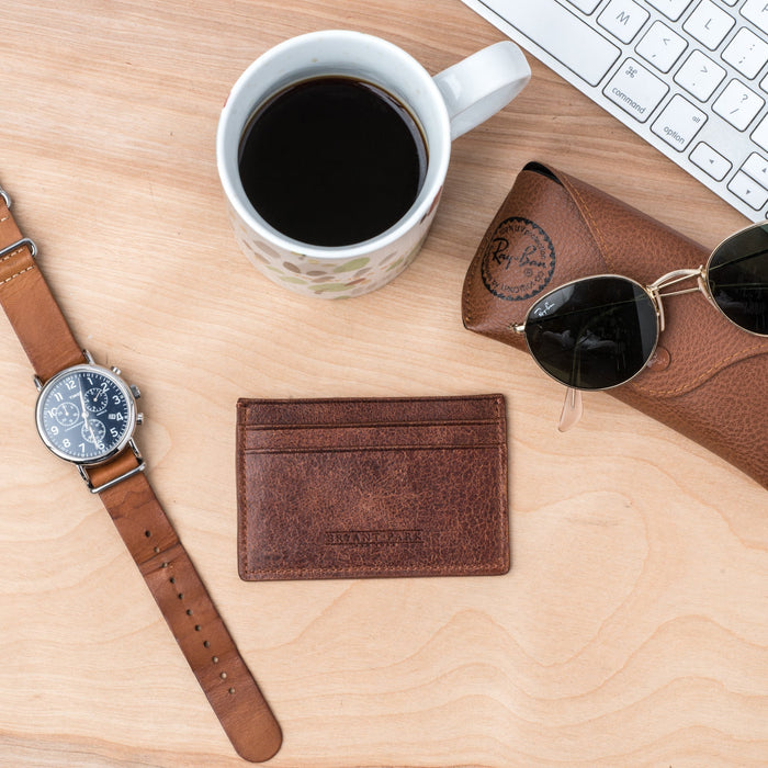 Brown leather watch, wallet, and sunglasses on a wooden desk with a cup of coffee and keyboard.
