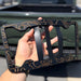 Person holding a decorative black metal license plate frame with a blurred green truck in the background.