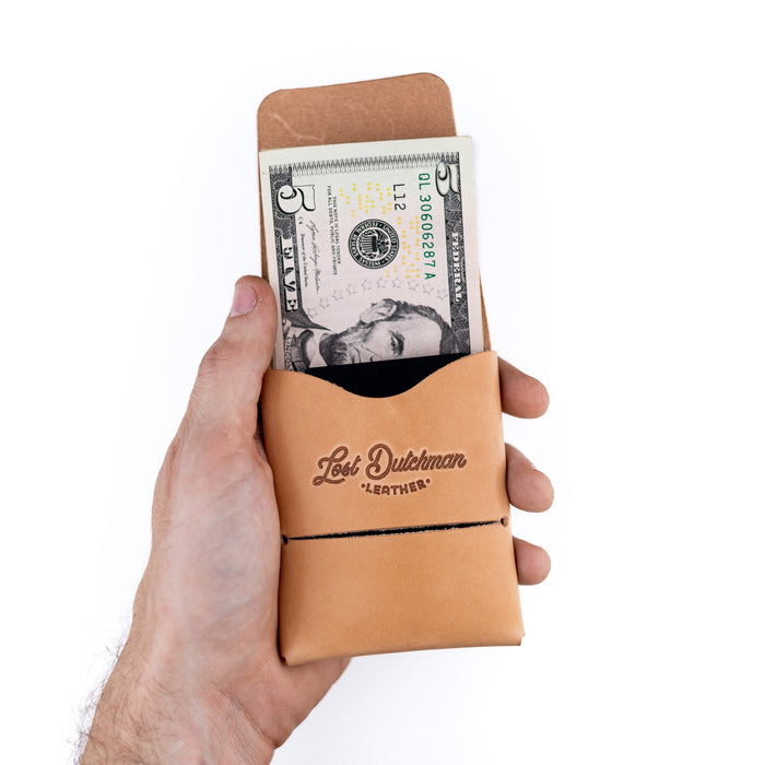 Hand holding a tan leather money clip with a dollar bill, branded 'Lost Dutchman Leather', on a white background.