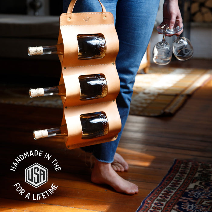 Person holding a wooden bottle holder with bottles and glasses indoors.
